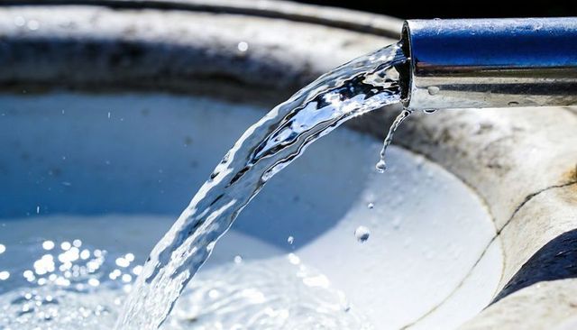 Stainless spout pouring clear water arc into sunlit stone basin with falling droplets