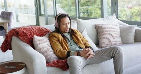 African american man relaxing on sofa wearing headphones listening to music in sunlit contemporary l
