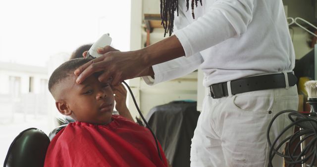 Young boy sitting in barbershop chair wearing red cape while barber with long hair is cutting his hair. This image underscores the skills and techniques employed in grooming children, highlighting both the concentration of the boy and the dexterity of the barber. Perfect for content related to hairdressing services, child grooming, family life, and community-oriented stories.