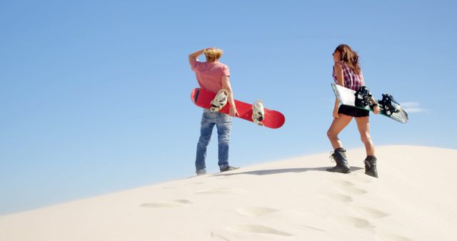 Caucasian couple carrying snowboards on a sand dune, with copy space. They're enjoying a unique outdoor adventure blending winter sports with a desert setting.