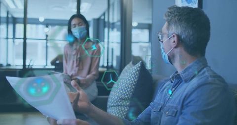 Masked colleagues collaborating over holographic data in modern office lounge