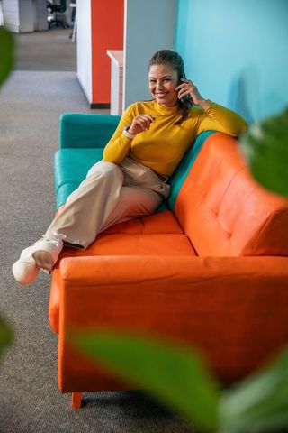 Smiling professional woman talking on phone while relaxing on vibrant office sofa