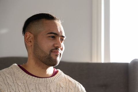Sunlit thoughtful man wearing cream cable-knit sweater sitting by window on gray couch