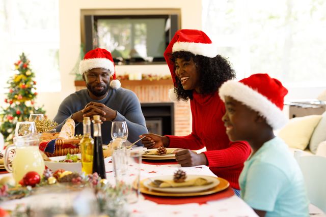 Front view of an african-american family enjoying their christmas dinner all wearing santa hats. a man can be seen on the edge of the table and a woman smiling beside him.