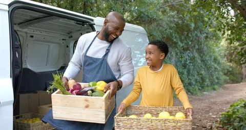 Father and son carrying fresh farm produce from white van, smiling, unloading crates on rural lane