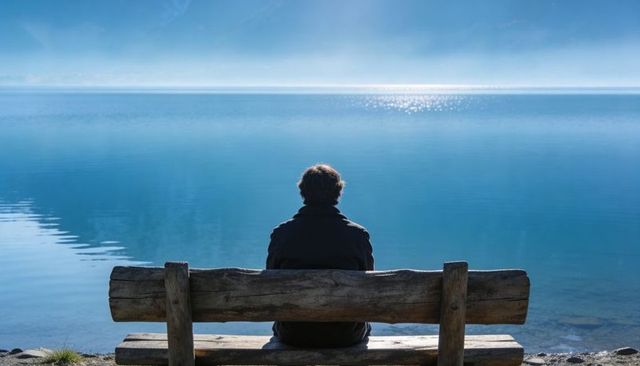 Solitary figure gazing over tranquil blue lake from weathered bench on rocky shore