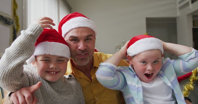 Portrait of caucasian father and two sons wearing santa hats waving and smiling sitting on the couch. christmas festivity and celebration concept