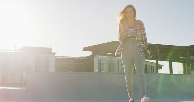 Young woman standing at a skate park holding a smartphone and a skateboard. She is dressed in casual wear and appears to be checking her phone. The environment is bright and sunny, suggesting a beautiful day for outdoor activities. This image can be used for themes related to youth culture, outdoor sports, lifestyle, and technology.