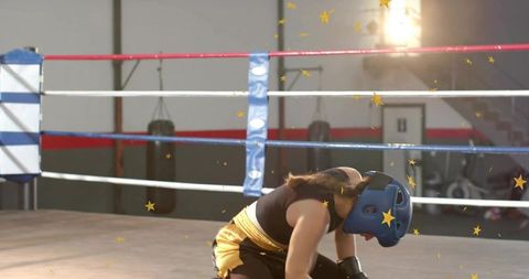 Kneeling female boxer catching breath in ring wearing blue headgear and yellow shorts