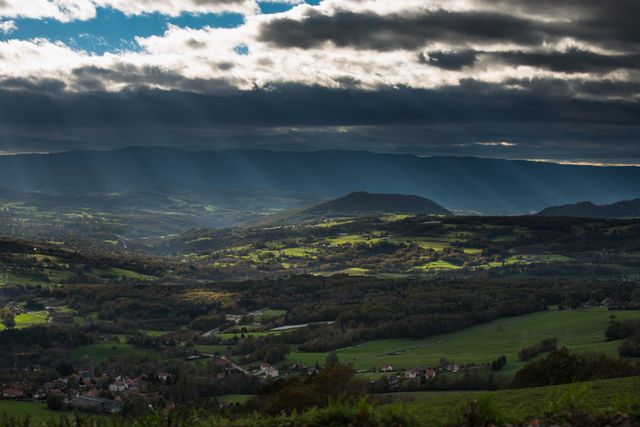 Dramatic scene of sunrays breaking through clouds, illuminating green valley surrounded by hills. Perfect for travel advertisements, nature magazines, environmental campaigns, backgrounds for inspirational quotes, and scenic posters.