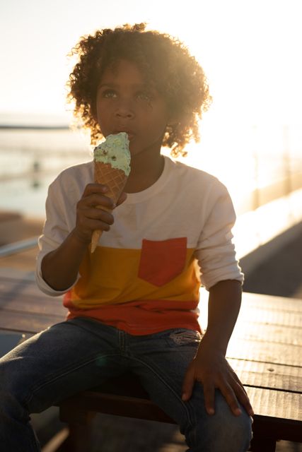 Front view of a biracial boy sitting on a bench enjoying an ice cream on a sunny day by the sea, backlit by the sun