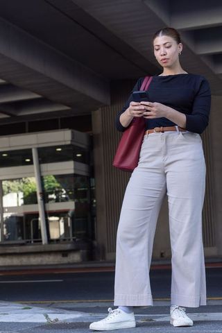 African american woman standing on curb checking smartphone, carrying red tote in city