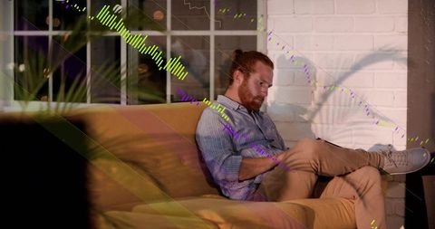 Redheaded man sitting on mustard sofa writing on tablet in cozy evening living room