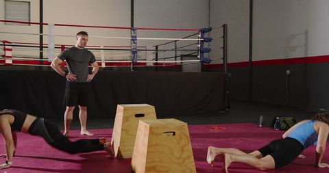 Trainer supervising female athletes during floor circuit and plyometrics near boxing ring