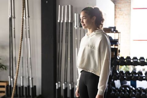Young woman standing in industrial gym wearing cropped hoodie and black leggings, sunlit