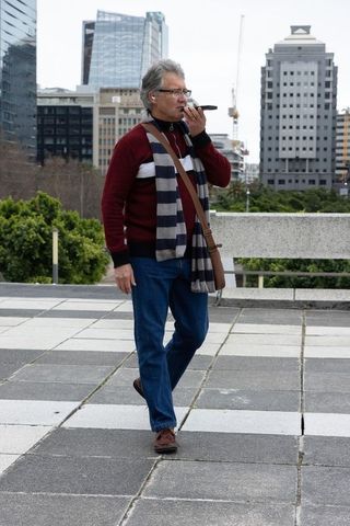 Senior man walking downtown plaza holding vape wearing striped scarf carrying brown bag