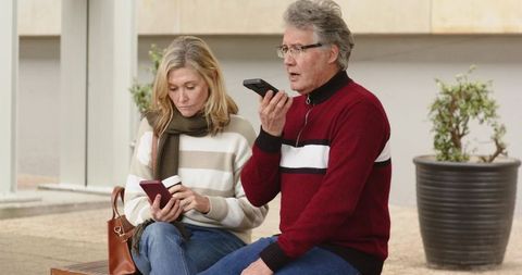 Mature couple sitting on wooden bench using smartphones while woman holding coffee cup