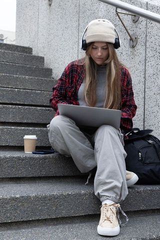 College student working on laptop sitting on concrete steps wearing beanie and headphones
