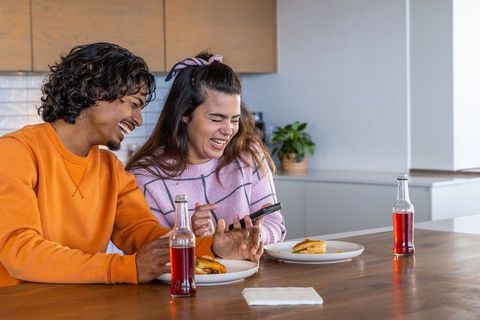 Diverse couple laughing and sharing sandwiches at modern kitchen island, casual moment