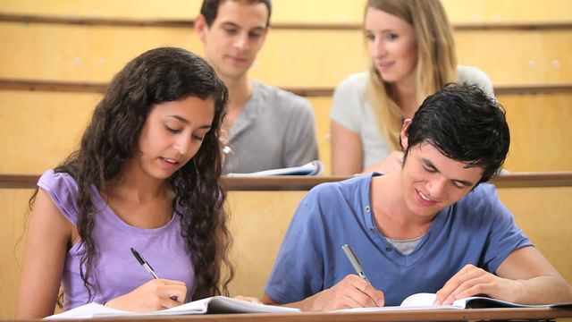 Group of university students sitting in a large lecture hall, actively engaged in note-taking during a class. They appear focused and collaborative, using notebooks and pens. Suitable for illustrating educational environments, group study scenes, or college experiences. Useful for materials on academic life, learning methods, or higher education promotions.