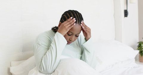 Mid-adult african american woman sitting on bed holding temples in light pajamas showing stress