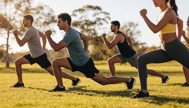 Male-led outdoor fitness class performing forward lunges on park grass at sunset