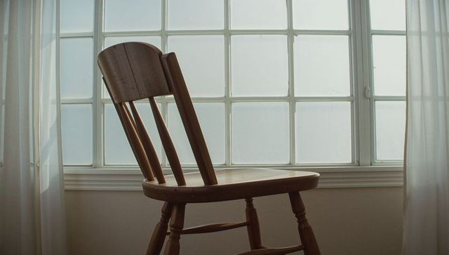 Sunlit wooden spindle-back chair leaning at multi-pane frosted window with sheer curtains