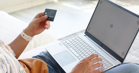 Asian man making online payment using credit card and laptop on sofa in bright daylight