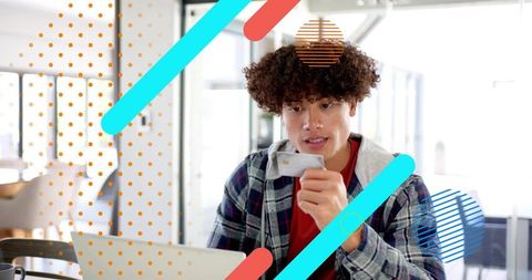 Curly young man holding phone using laptop in modern coworking space with colorful overlays