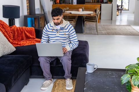 Indian man working from home on laptop on modern sectional sofa in sunlit living room