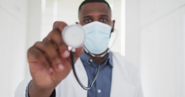 African American doctor holds stethoscope towards camera while wearing face mask in a bright medical office. Ideal for healthcare, medical practice, pandemic safety, and patient care materials.