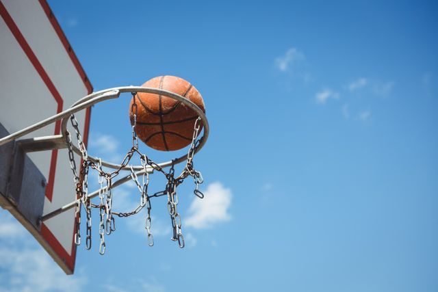 This image captures a basketball going through an outdoor hoop with a chain net against a clear blue sky. Ideal for use in sports-related content, motivational posters, recreational activity promotions, or articles about outdoor sports and fitness.