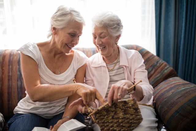 Cheerful senior women talking about knitting while sitting on sofa against window at retirement home