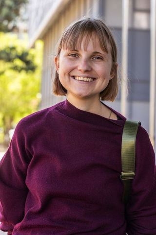 Smiling young woman on city sidewalk sporting maroon sweater and green backpack strap
