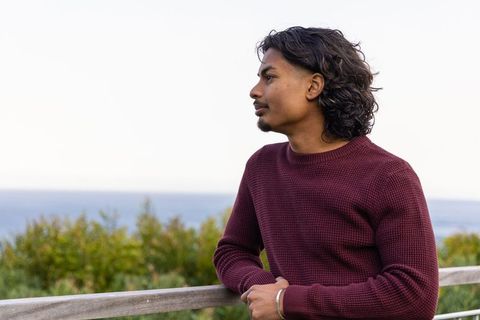 Indian man leaning on boardwalk railing at seaside wearing maroon sweater contemplative