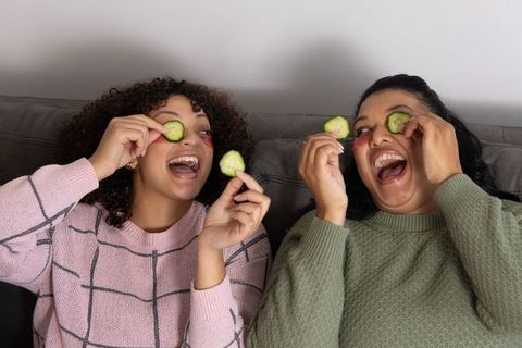 Friends laughing holding cucumber eye slices and gel patches on couch for at-home skincare