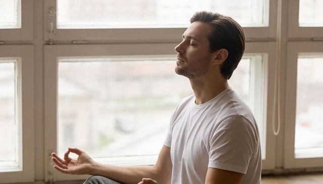 Young man meditating by large window in minimalist room, mindfulness and relaxation