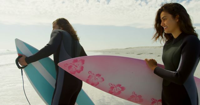 Two women standing on a sunny beach holding colorful surfboards, preparing for surfing in the ocean. They are wearing wetsuits and appear to be enjoying their time outdoors. This image can be used for promoting sports, outdoor activities, beach lifestyle, summer vacations, and fitness.