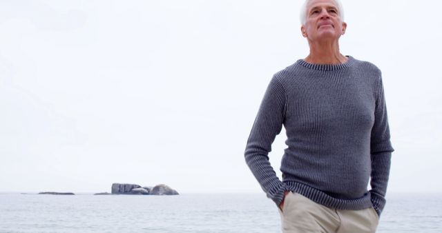 Elderly man standing near ocean in grey sweater, hands in pockets, looking contemplative. Use this image in health and wellness, retirement planning, leisure activities, blogs, or lifestyle articles highlighting peaceful living and reflection.