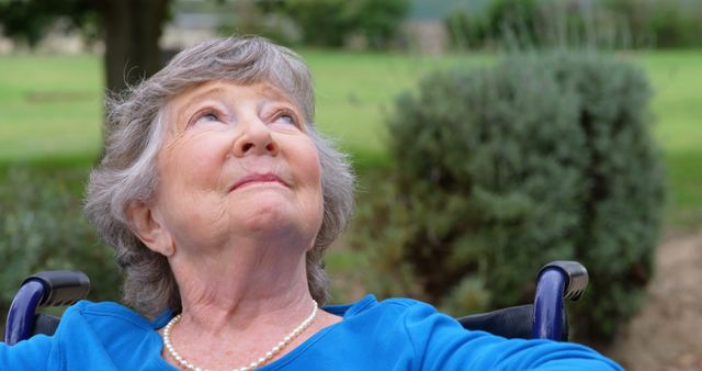 The photo captures a senior woman sitting in a wheelchair, smiling and looking up, suggesting contemplation or enjoyment of nature. Ideal for depicting positive aging, health care services, elderly independence, and outdoor activities. Can be used in advertising for retirement communities, medical care for elderly, or lifestyle magazines aimed at older audiences.