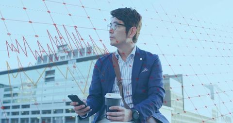 Asian businessman checking smartphone and holding coffee amid financial data overlay in urban plaza