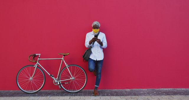 African american senior man wearing face mask with bicycle using smartphone while standing against pink wall. hygiene and social distancing during coronavirus covid-19 pandemic.