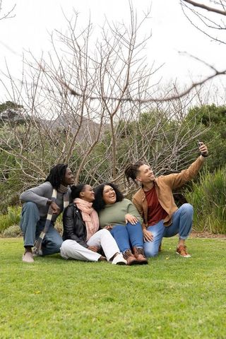 Multiracial friends laughing and taking selfie on green lawn during casual outdoor meetup