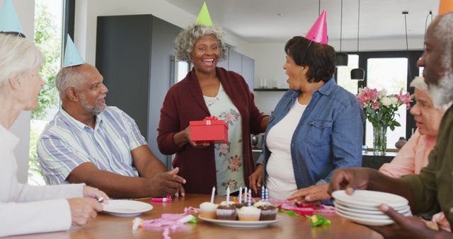 Senior friends enjoying a birthday celebration together, smiling with party hats, exchanging a gift, and preparing to eat cake with candles. Ideal for use in content related to elderly social events, community involvement, and the joy of aging with friends.