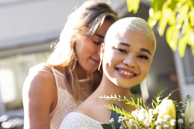 Multiracial young woman with eyes closed cuddling girlfriend with short hair from behind in yard. Happy, unaltered, love, togetherness, homosexual, romance and lifestyle concept.