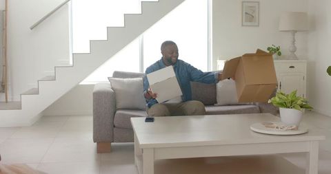 African american man unpacking moving boxes on sofa in bright modern living room daylight