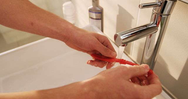 Close-up view of hands washing a red toothbrush under running water in a modern bathroom sink. Highlights good dental hygiene and daily cleanliness routine. Useful for dental health blogs, morning routine articles, cleanliness campaigns, and personal hygiene promotions.