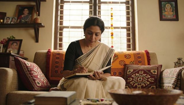 Middle-aged indian woman writing journal on cozy sofa with traditional woven textiles