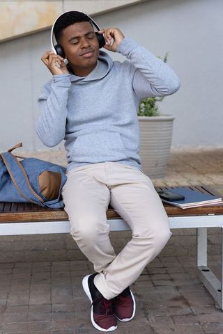 African american teen relaxing on bench listening to music with headphones and backpack
