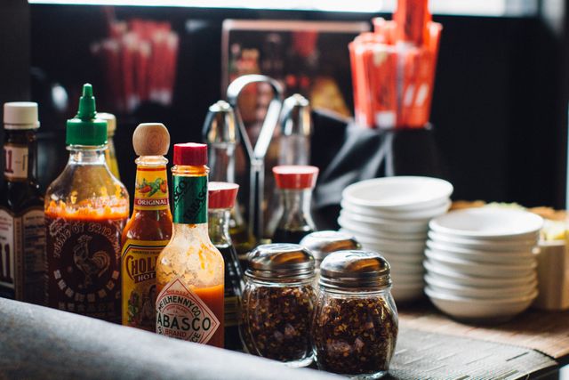 Assorted sauce bottles including hot sauces, soy sauce, and mixed seasoning jars on a restaurant table. Nearby are several clean, stacked white dishes. Ideal for use in food service promotions, restaurant advertisements, or culinary blog posts showcasing dining atmosphere.